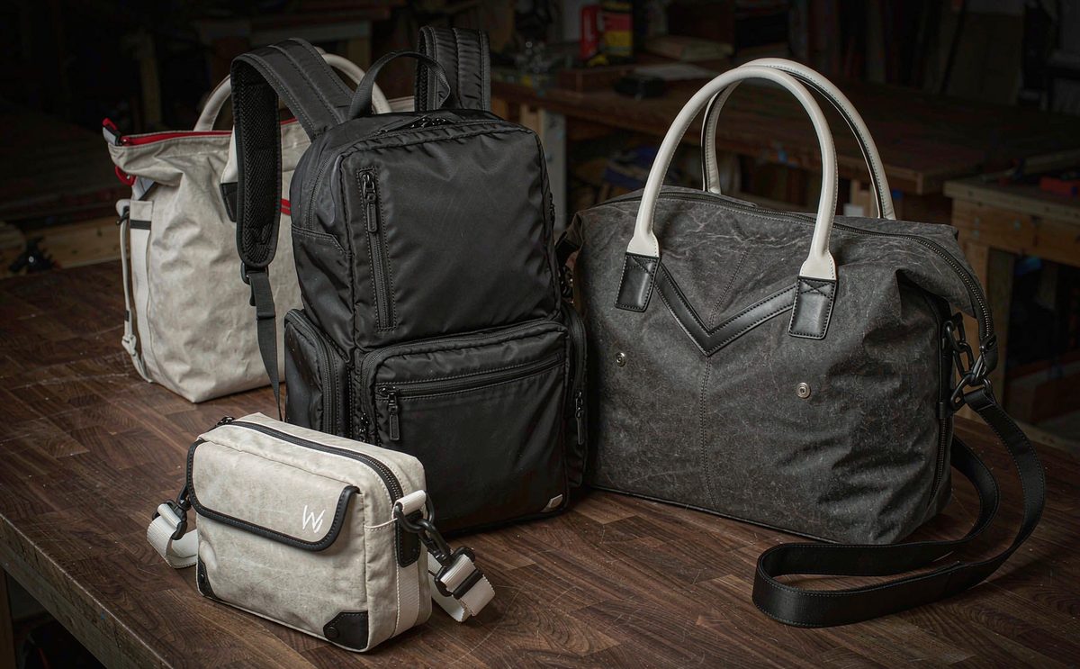 A black backpack, a charcoal duffel, a waxed canvas tote and a zippered pouch on a workbench