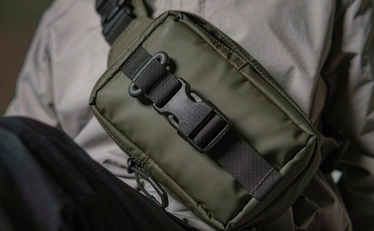 A compact olive crossbody bag with an adjustable webbing strap worn across a grey jacket