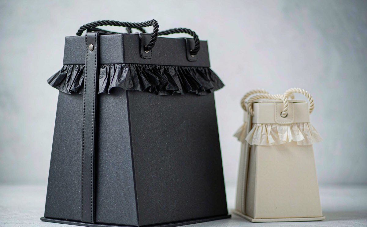 A matte black gift bag with rope handles standing beside a smaller cream one